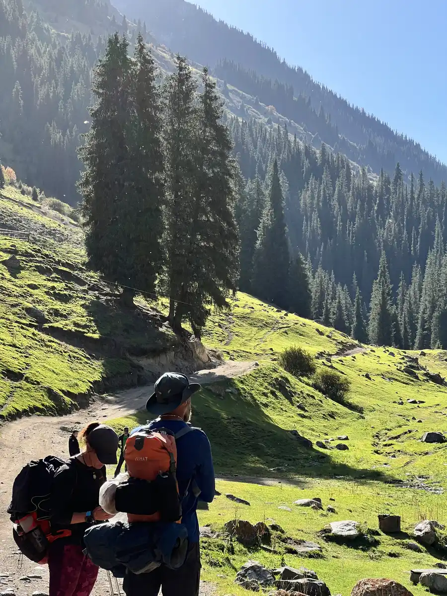 Two backpackers on a trail through a green pine-forested Kyrgyz mountain valley